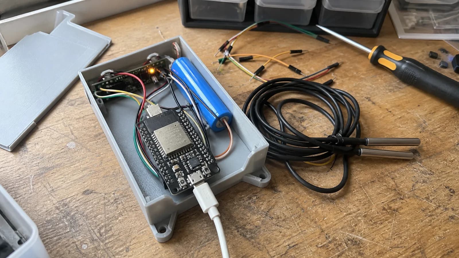 Prototype thiết bị báo nhiệt độ tủ lạnh gồm board ESP32, pin dự phòng nhỏ, hộp in 3D và hai đầu dò DS18B20 chống nước đặt trên bàn làm việc.