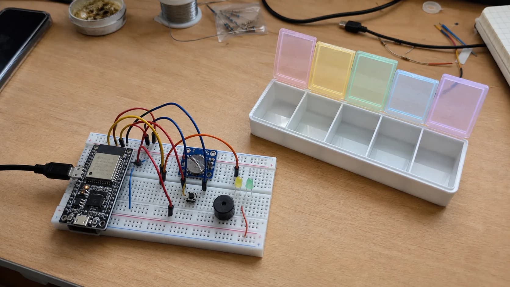 Prototype hộp nhắc uống thuốc dùng ESP32, module RTC, buzzer, LED và hộp thuốc nhiều ngăn trên bàn làm việc.